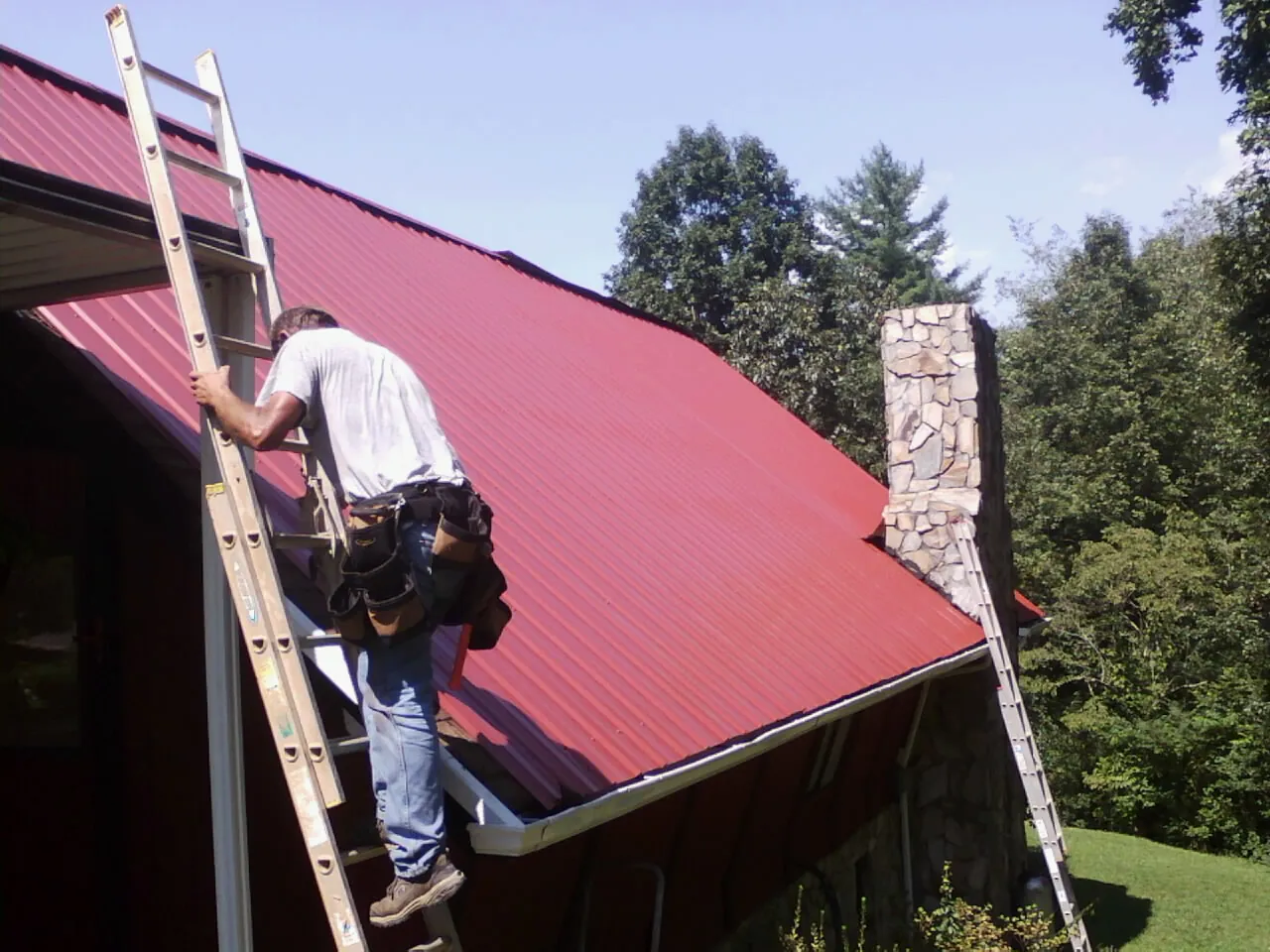Roofer on a ladder beside a red metal roof and stone chimney — Soffit & Fascia Repair by Roofing Brothers in Emeryville